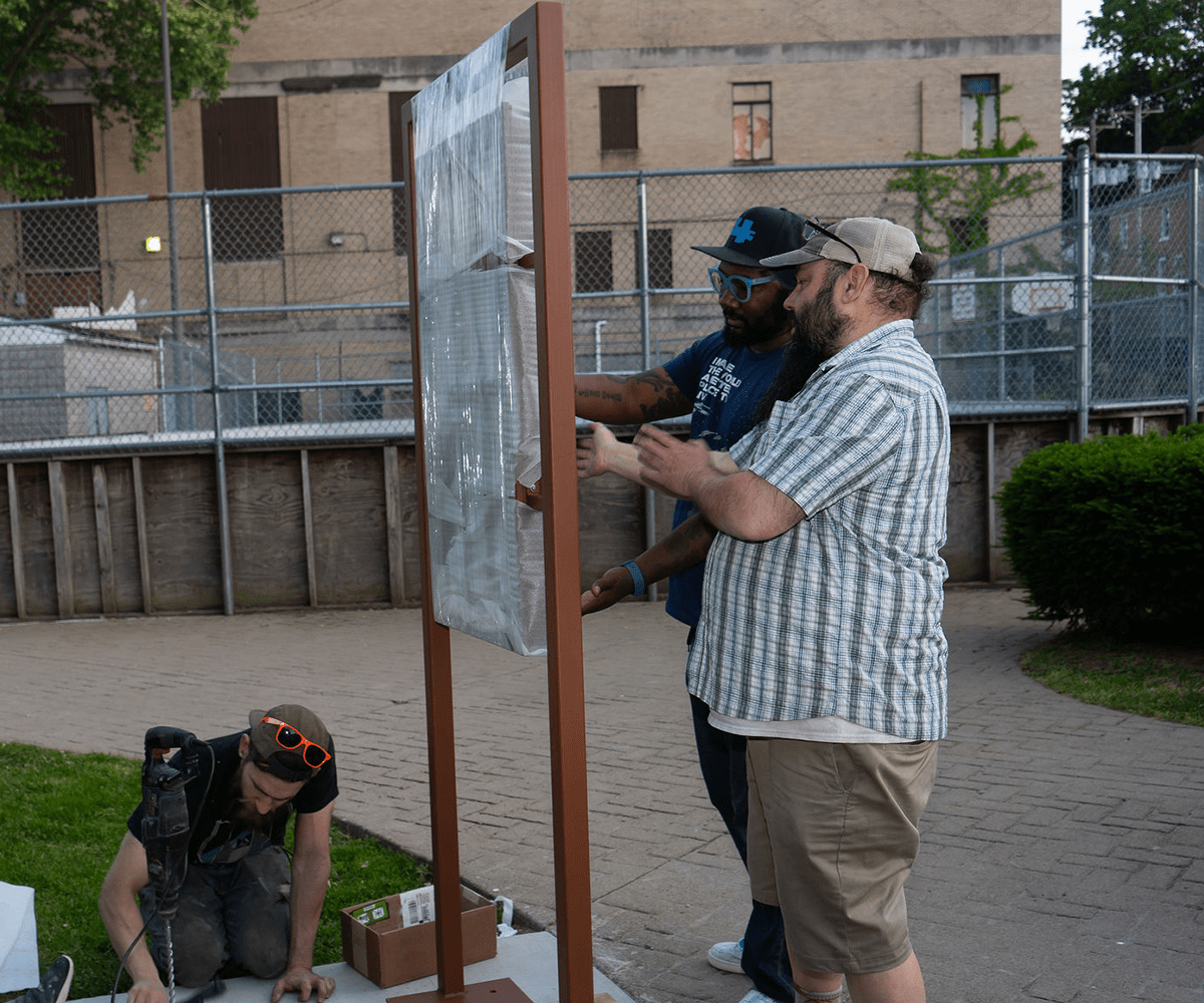 Three people, including artist Jason McKoy, are working on installing a steel frame onto a concrete slab.