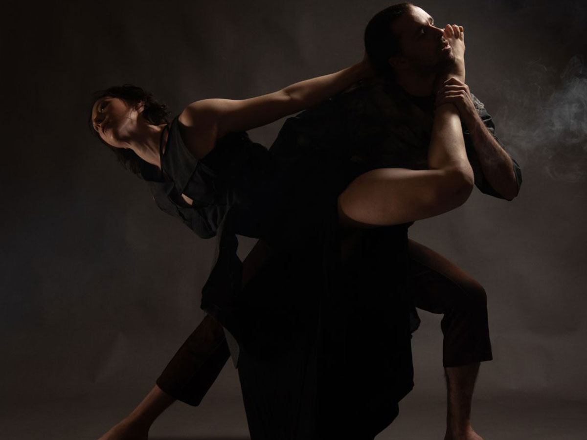 two dancers are intertwined in a studio shoot in front of a gray backdrop. One dance is facing the left, arching up towards the light as the other dancer faces the right, bringing the foot of the other dancer up towards their face.