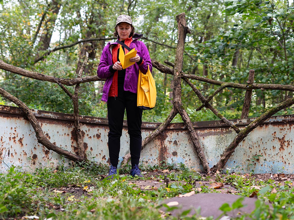 Erin Mallea is standing amidst a forest and an aging metal structure. She is reading aloud from a yellow booklet that she holds in her hands. She is wearing a purple jacket, black pants, and a baseball hat. A yellow tote is slung in her left elbow.