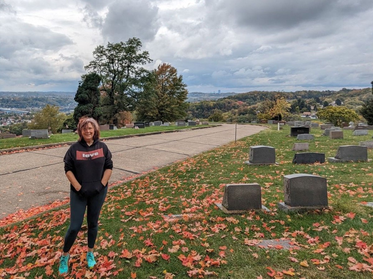 Poet Nancy Krygowski is standing in a cemetery. It is autumn, and there are orange leaves on the ground.