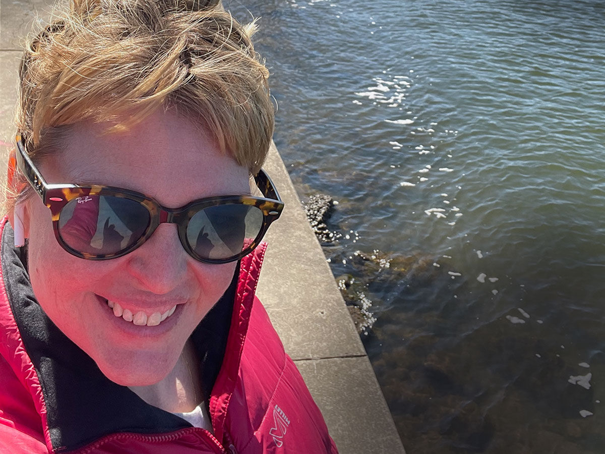 Sarah Shotland is standing next to a river wearing a red puffy jacket and sunglasses.