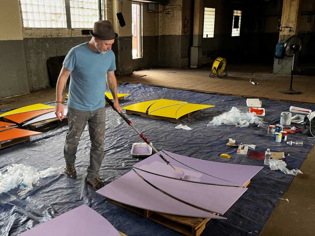 Artist Tim Engelhardt is in a large warehouse working on elements of the mural for Pittsburgh CLO. In this photo, the piece he is working on is on the ground on top of large plastic sheeting spread across the floor. Tim is using purple paint and a long handled paint roller.