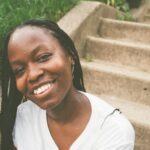 Maya Marie S. is sitting on an outdoor set of concrete stairs. She is wearing a white tee-shirt and smiling. 