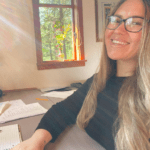 Christina Berke is seated at a desk with papers on it. There is sunlight streaming through a window. She is wearing a black long-sleeved top and glasses. 