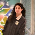 Max Gonzales is standing in front of a graffiti artwork, leaning against a chain-link fence. 