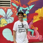 Shane Pilster is photographed in front of a graffitied wall. He is wearing a white tee-shirt with a graphic on it that reads: Sobriquets, Monikers, Aliases & Epithets.