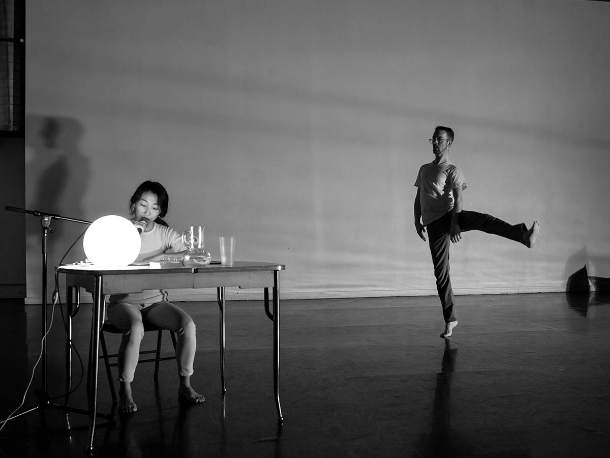 Black and white image of a man dancing in the middle of a room. He has his left leg raised, while standing on the toes of his right foot; his hands are at his sides. Another person is in the space, seated at a desk with a glowing orb light on it. They are speaking into a microphone.