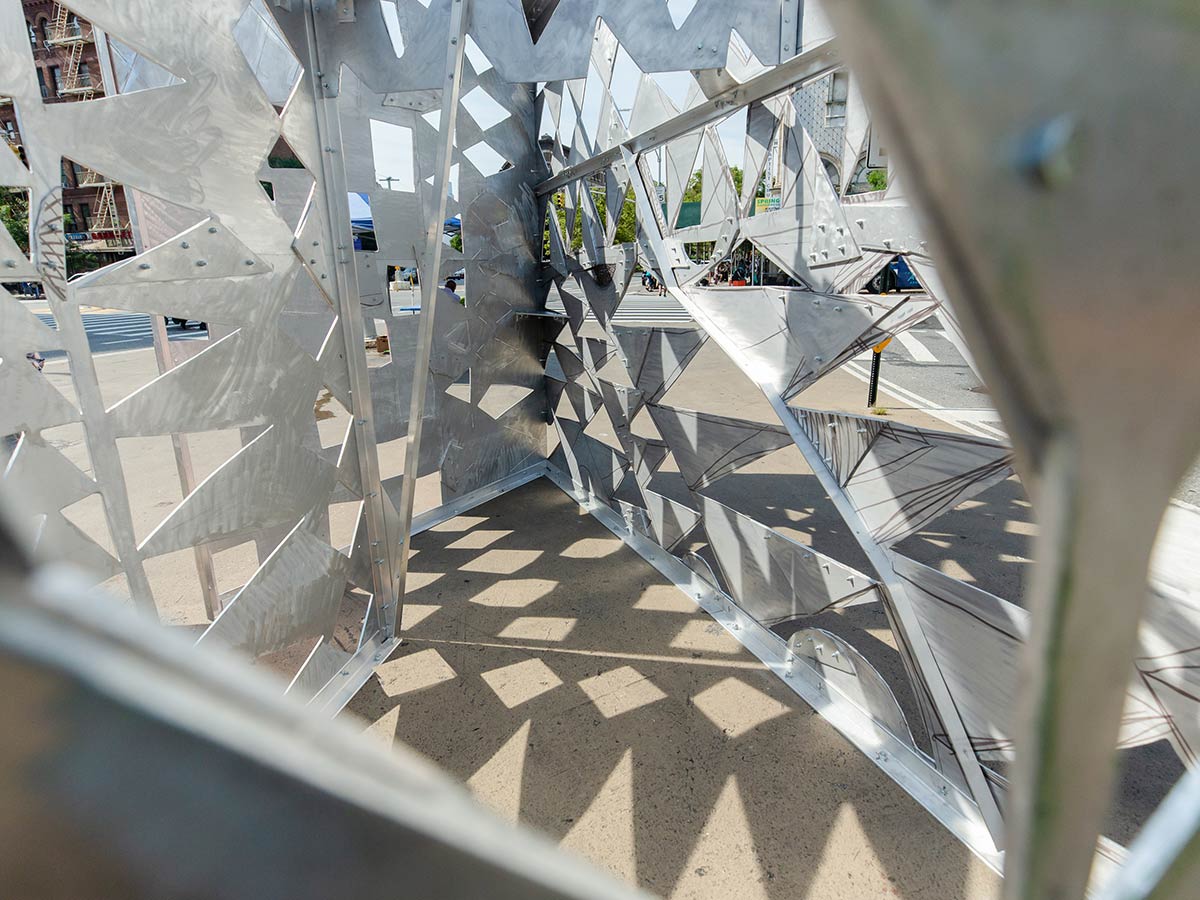 A metal sculpture is outdoors on a concrete island in an urban setting. It has a repeating series of triangular shapes cut out of it, creating a patterned shadow on the concrete. The interior of the sculpture is silver.