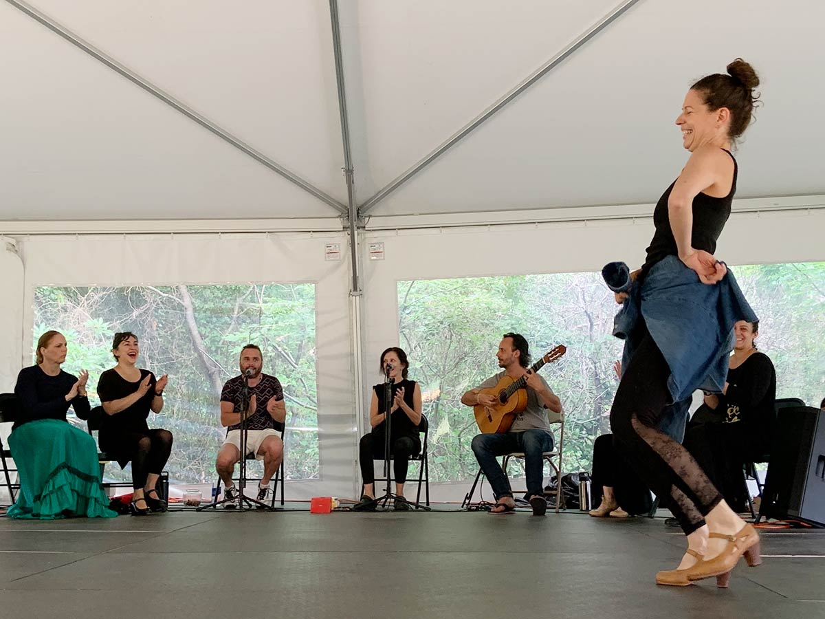 Leslie Roybal is inside a white tent. She is dressed in dance attire and is photographed mid-dance step. There are musicians and singers seated in the background.