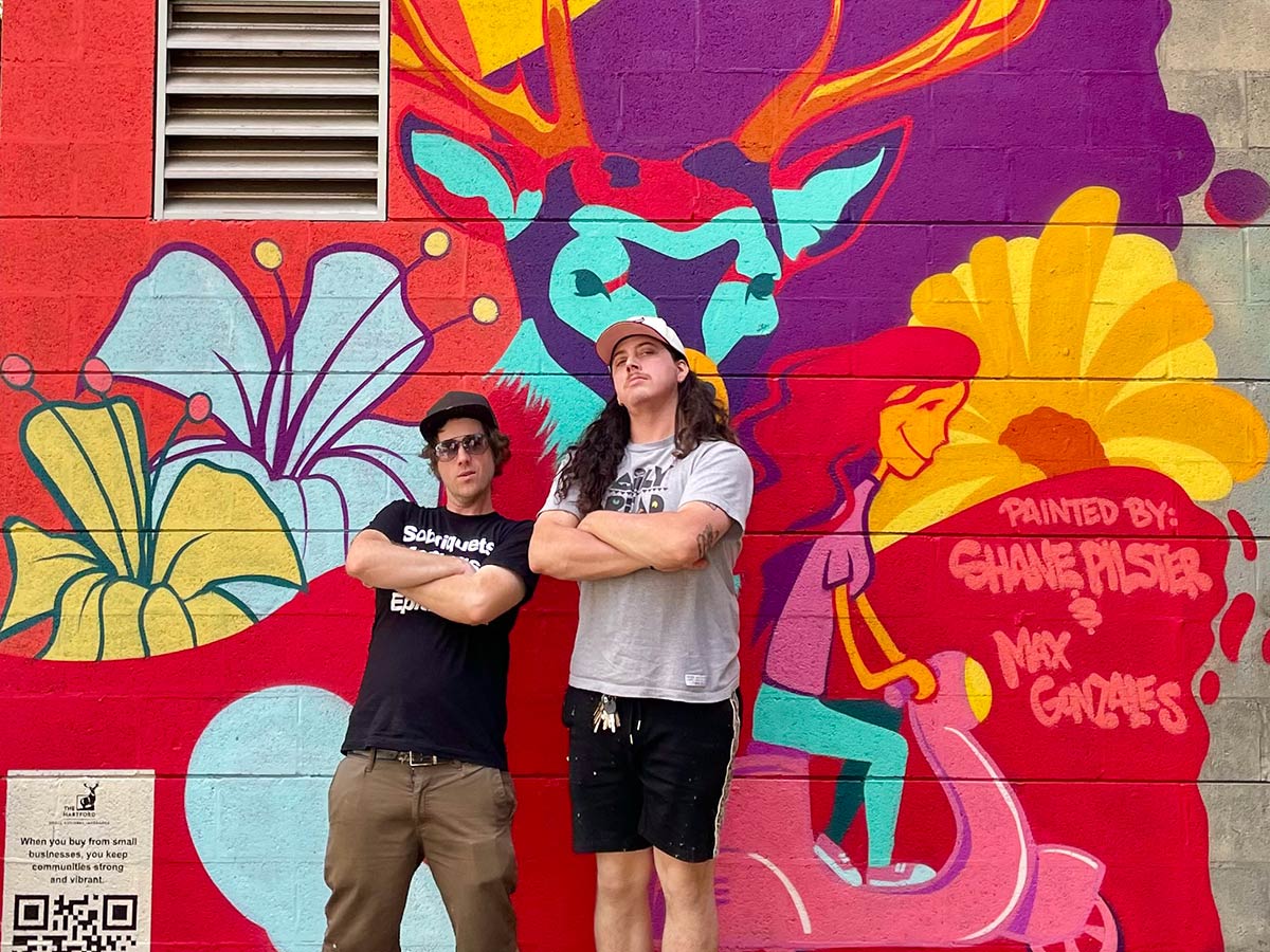 Max Gonzales (right) and Shane Pilster (left) stand against one of their murals. It is in bold shades of red, purple, light blue, golden yellow, teal, and pink.