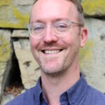 David Bernabo is standing against a stone wall, wearing glasses and a blue collared shirt.