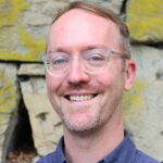 David Bernabo is standing against a stone wall, wearing glasses and a blue collared shirt.