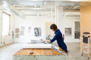 Artist Kim Beck is wearing a navy blue dress, working on a project at a waist-high wood table. She is in an industrial-style studio with artwork on white walls in the background.