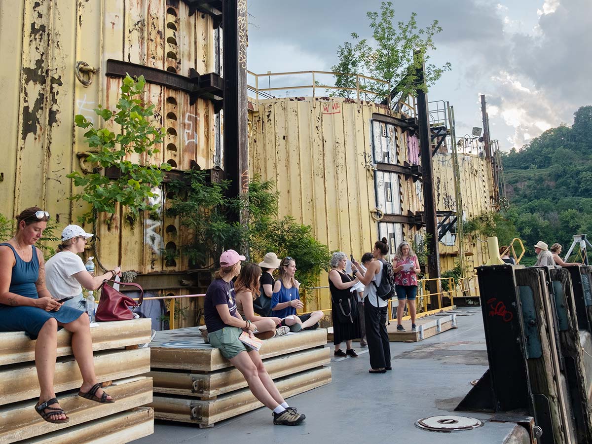 Several people are seated and standing around an industrial dock along the Monongahela River in Hazelwood Green. They are all observing and writing.